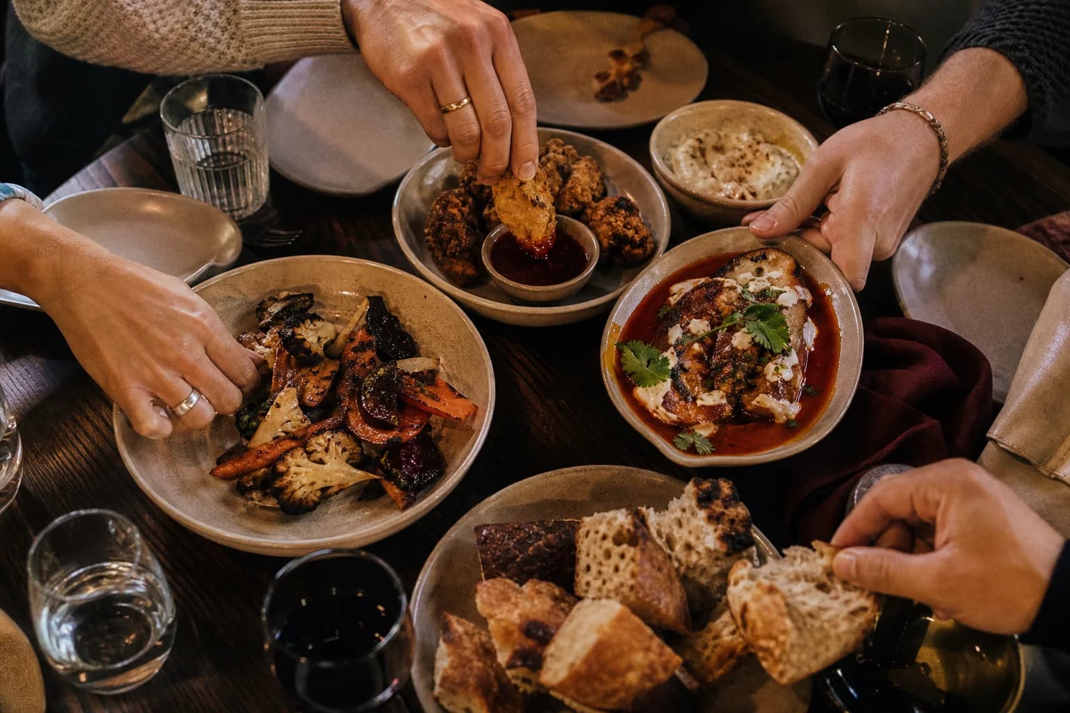 Friends share warm plates, bread, and roasted vegetables around a dark table.